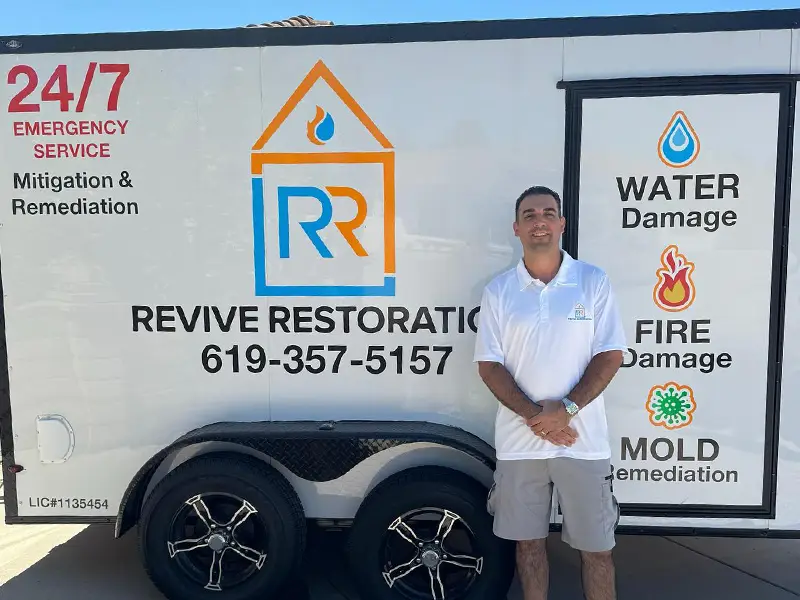 Man in white polo and shorts stands beside a trailer advertising 24/7 emergency water, fire, and mold damage restoration. Working for a water damage restoration in San Diego company