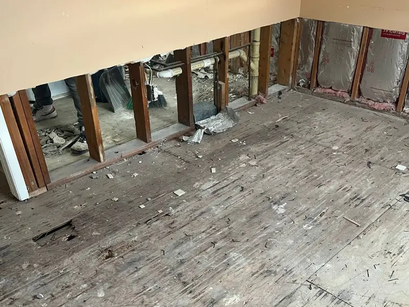 Room under renovation with exposed wall studs and a worn wooden floor covered in debris.